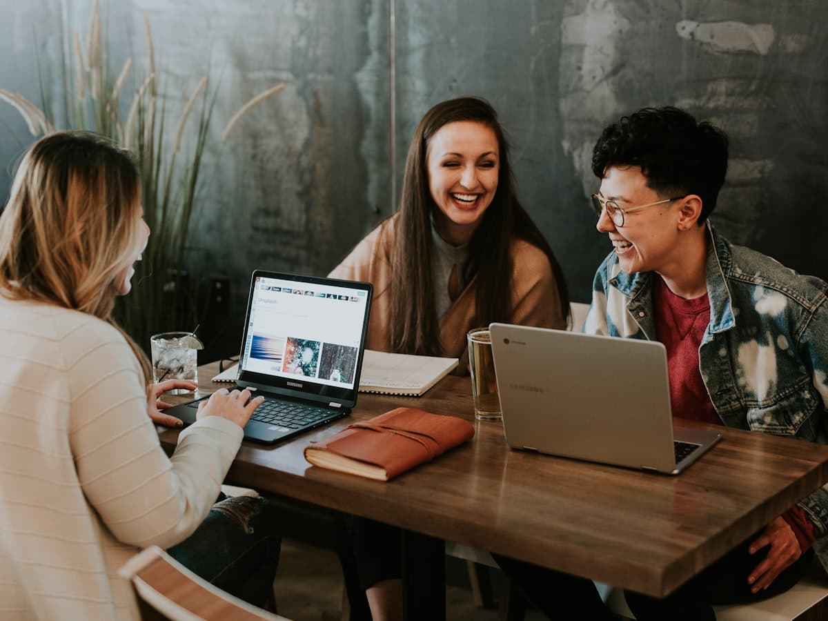 Three learners laughing together while collaborating on laptops around a shared table